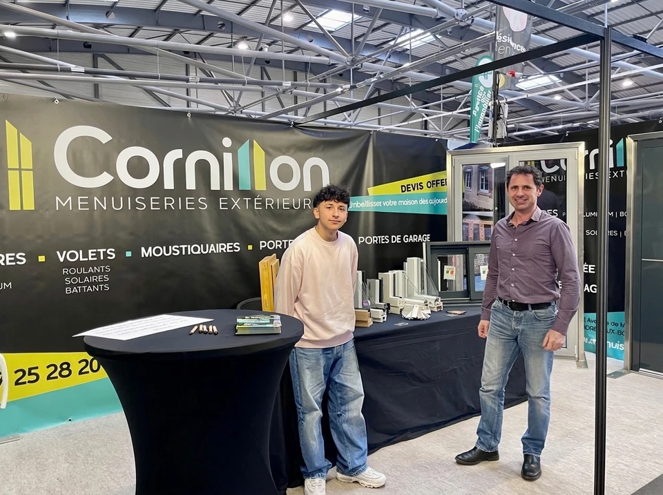 Stand d'exposition professionnel en intérieur, deux hommes debout devant un panneau publicitaire noir de l'entreprise **Cornillon Menuiseries Extérieures**